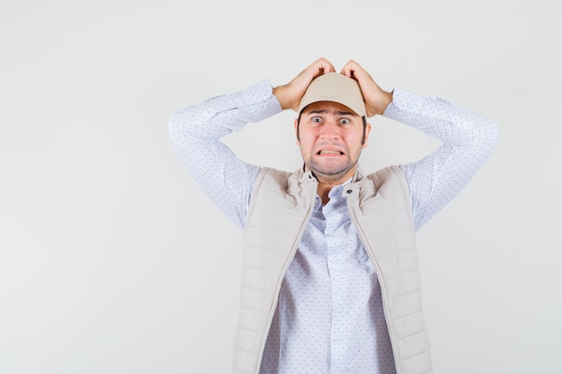 Young man holding hands on head while panicking in shirt,sleeveless jacket,cap and looking anxious. front view.