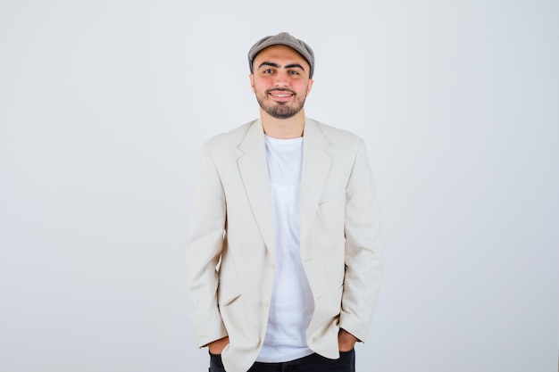 Young man holding hands in pocket in white t-shirt, jacket and grey cap and looking happy