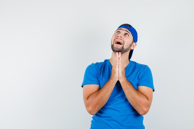 Young man holding hands in praying gesture in blue t-shirt and cap and looking hopeful