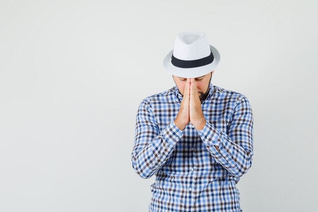 Young man holding hands in praying gesture in checked shirt, hat and looking calm. front view.