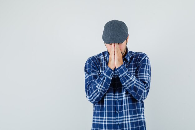 Молодой человек, взявшись за руки в молитвенном жесте в рубашке, кепке и выглядя обнадеживающим, вид спереди.