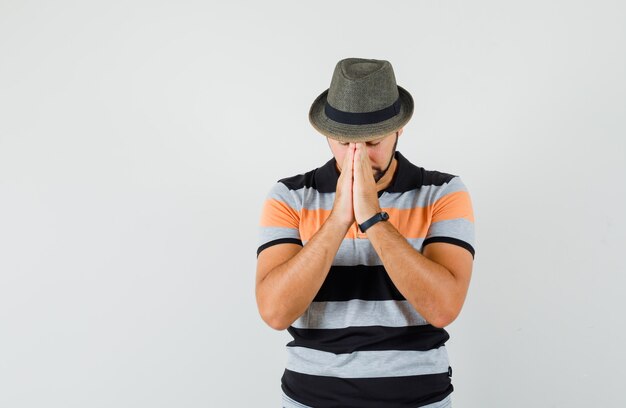 Молодой человек, взявшись за руки в молитвенном жесте в футболке, шляпе и глядя с надеждой, вид спереди.