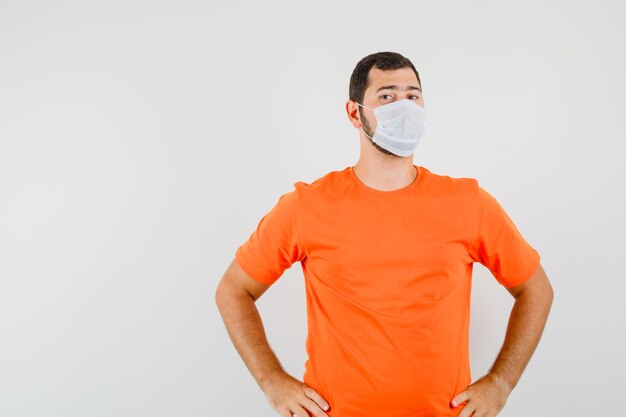 Young man holding hands on waist in orange t-shirt, mask and looking confident. front view.