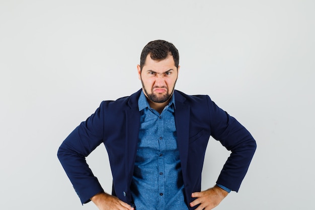 Free photo young man holding hands on waist in shirt, jacket and looking furious. front view.