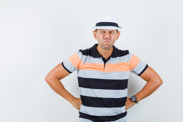 Free photo young man holding hands on waist in t-shirt, hat and looking sullen