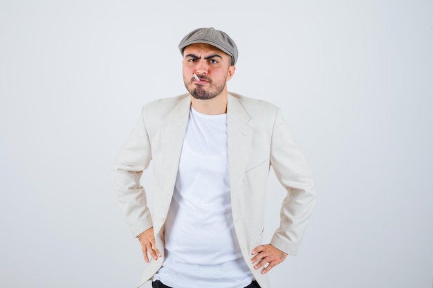 Young man holding hands on waist while smoking in white t-shirt, jacket and gray cap and looking angry