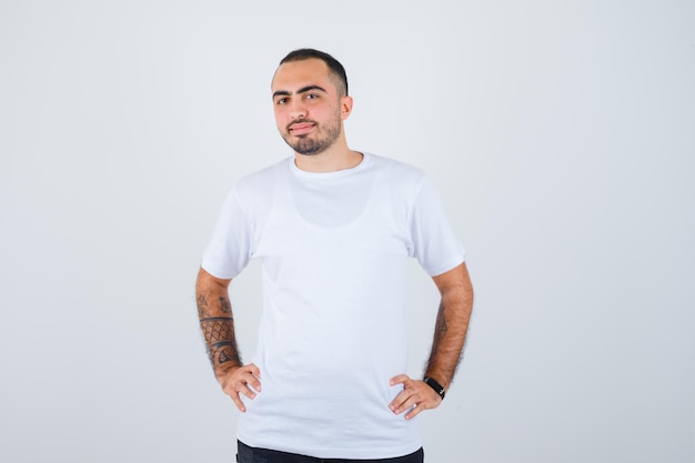 Young man holding hands on waist in white t-shirt and black pants and looking serious