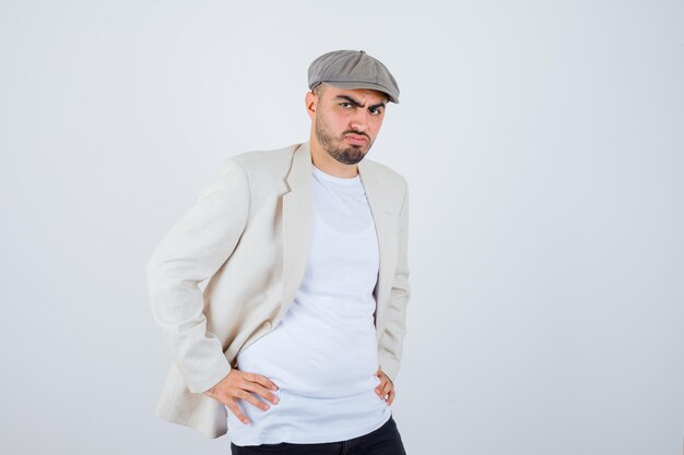 Young man holding hands on waist in white t-shirt, jacket and gray cap and looking serious