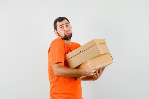 Young man holding heavy cardboard boxes in orange t-shirt , front view.