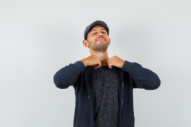 Young man holding his collar in t-shirt, jacket, cap and looking proud. front view.