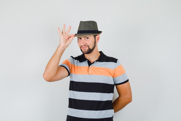 Молодой человек держит шляпу в футболке и выглядит мило, вид спереди.
