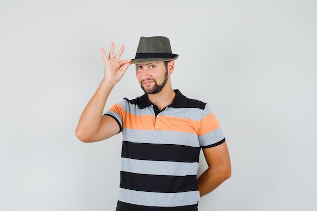 Young man holding his hat in t-shirt and looking cute , front view.