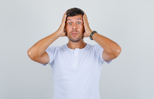 Young man holding his head on hands in white t-shirt and looking anxious, front view.