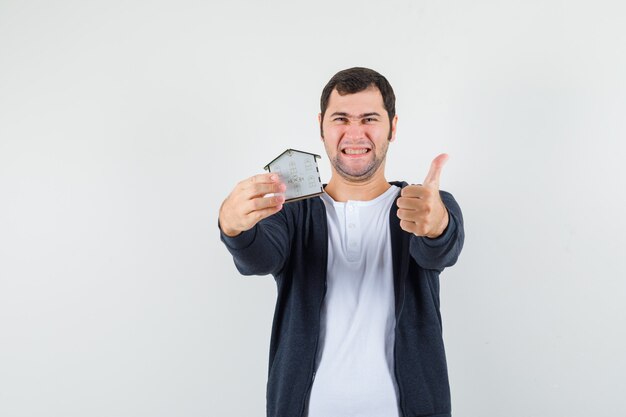 Young man holding house model in one hand and showing thumb up in white t-shirt and zip-front black hoodie and looking optimistic , front view.