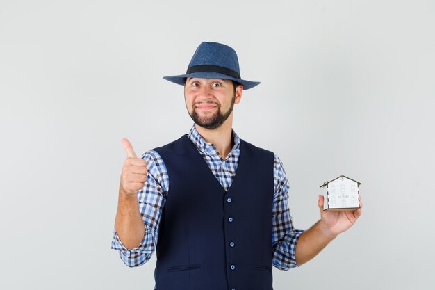 Young man holding house model, showing thumb up in shirt, vest, hat and looking merry , front view.