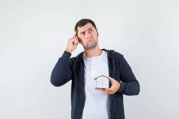 Young man holding house model, thinking about something in white t-shirt and zip-front black hoodie and looking pensive. front view.