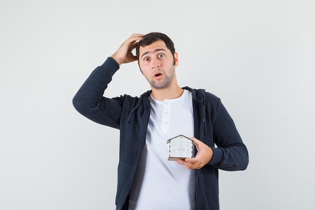 Young man holding house model, thinking about something in white t-shirt and zip-front black hoodie and looking surprised. front view.