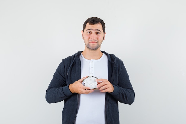 Young man holding house model with both hands in white t-shirt and zip-front black hoodie and looking serious , front view.