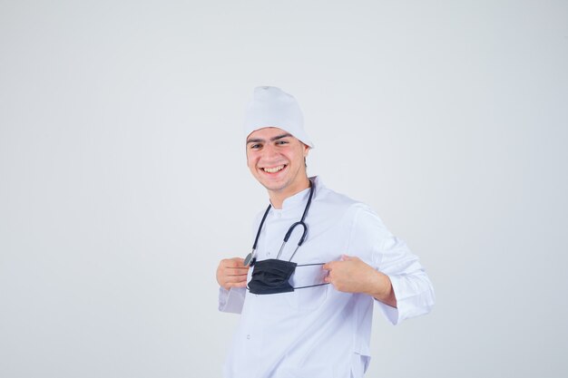 Young man holding mask while smiling in white uniform and looking cheerful. front view.