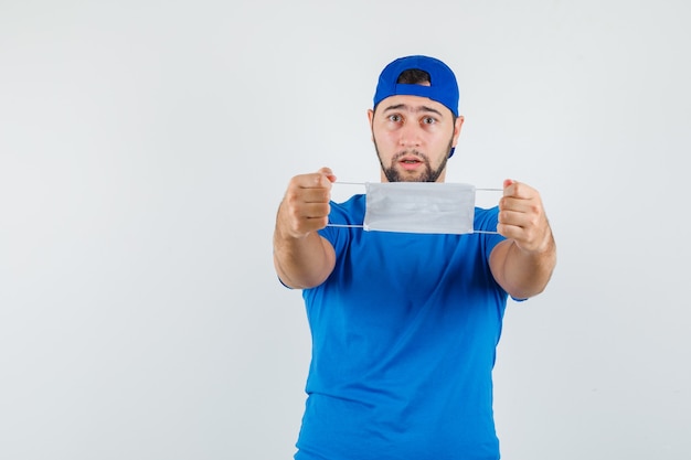 Young man holding medical mask in blue t-shirt and cap and looking careful 