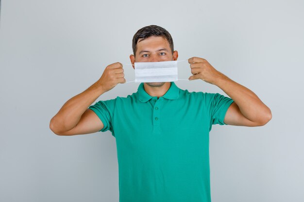 Young man holding medical mask over mouth in green t-shirt and looking careful, front view.