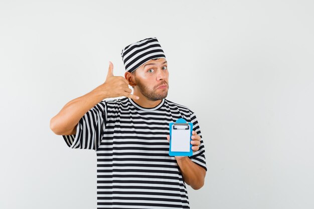 Young man holding mini clipboard showing phone gesture in striped t-shirt hat 