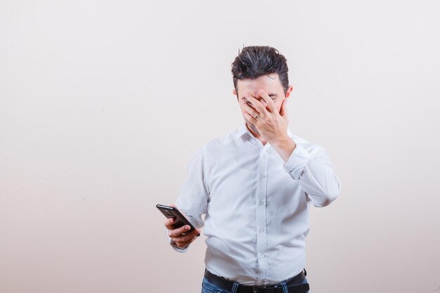 Young man holding mobile phone, covering face with hand in shirt, jeans and looking anxious