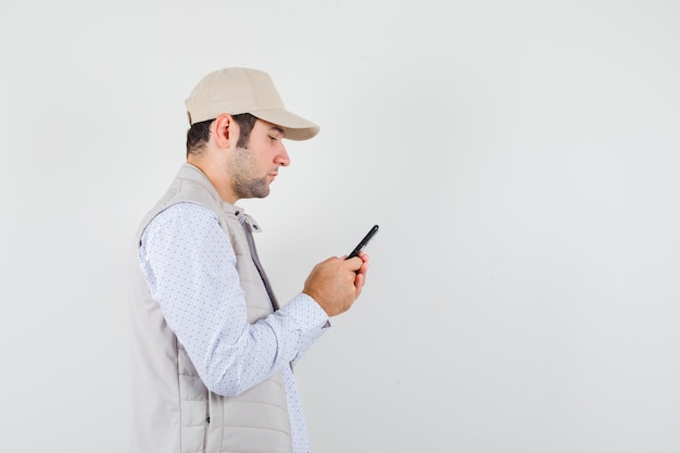 Young man holding mobile phone at hand in beige jacket and cap and looking serious. front view.