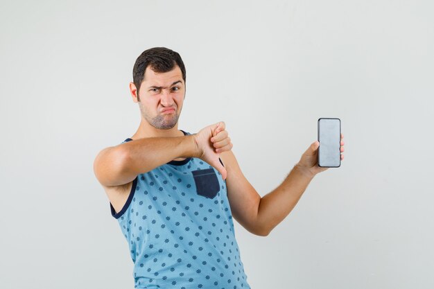 Young man holding mobile phone, showing thumb down in blue singlet and looking discontent