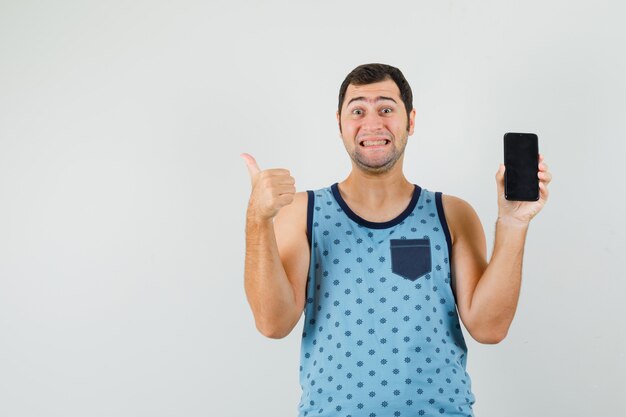 Young man holding mobile phone, showing thumb up in blue singlet and looking happy