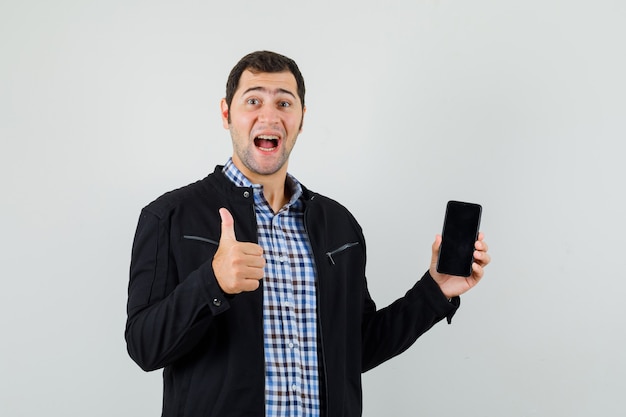 Young man holding mobile phone, showing thumb up in shirt, jacket and looking glad