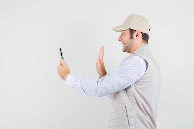 Young man holding mobile phone and talking someone via video call in beige jacket and cap and looking happy , front view.