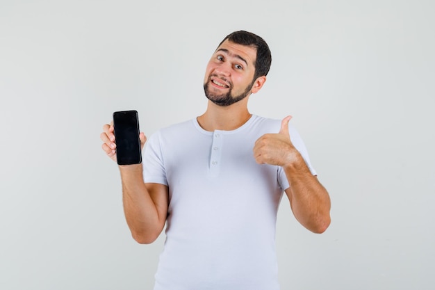 Young man holding mobile phone while showing thumb up in white t-shirt and looking happy. front view.