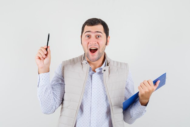 Young man holding notebook and pen while raising index finger in eureka gesture in beige jacket and looking excited. front view.