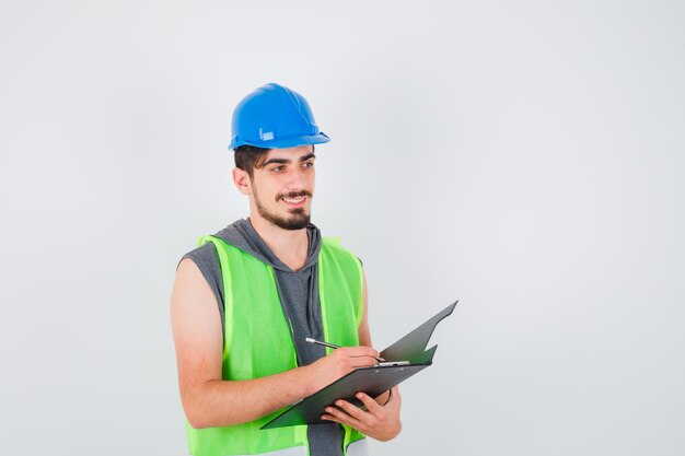 Young man holding notebook and writing something on it with pen in construction uniform and looking happy