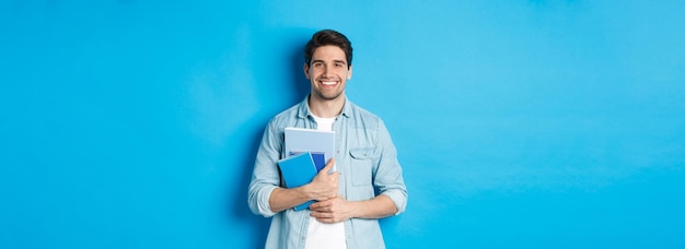 Бесплатное фото Молодой человек с блокнотами и учебным материалом улыбается, счастливо стоя на синем фоне