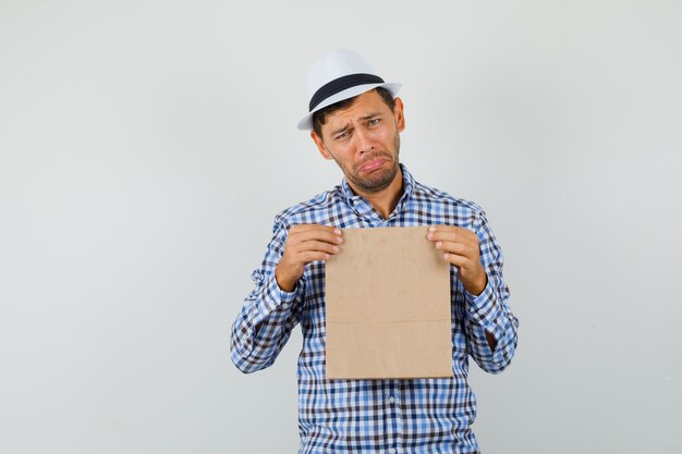 Free photo young man holding paper bag in checked shirt, hat and looking downcast