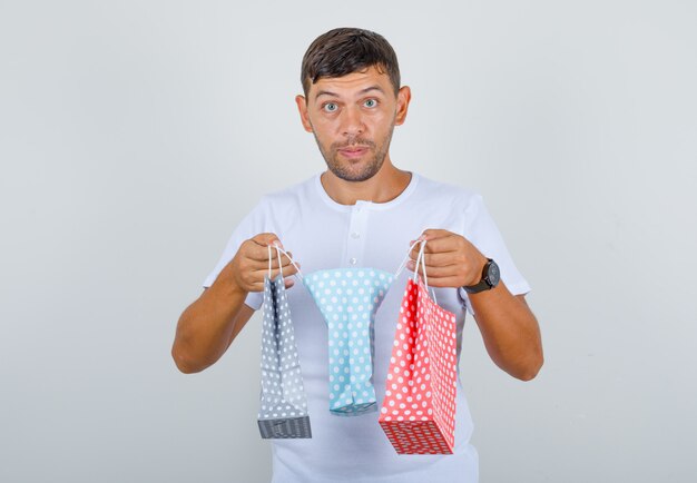 Young man holding paper bags in white t-shirt and looking confused, front view.