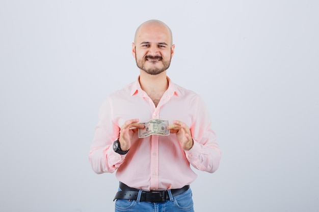 Молодой человек держит бумажные деньги в розовой рубашке, джинсах и выглядит довольным, вид спереди.
