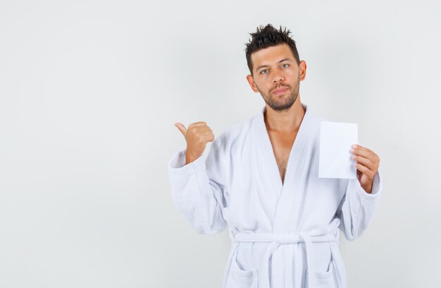 Young man holding paper sheet while pointing back in white bathrobe , front view.