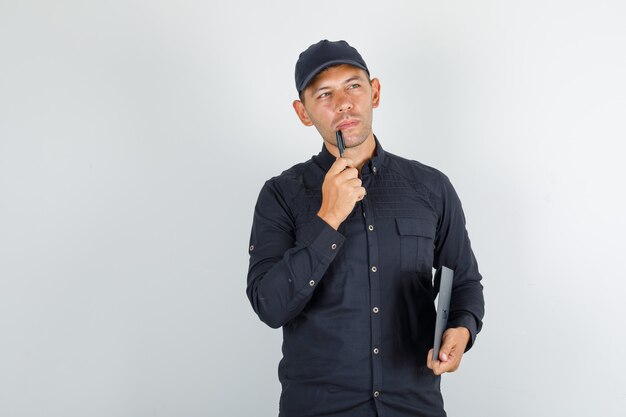 Young man holding pen and folder in black shirt and cap and looking thoughtful