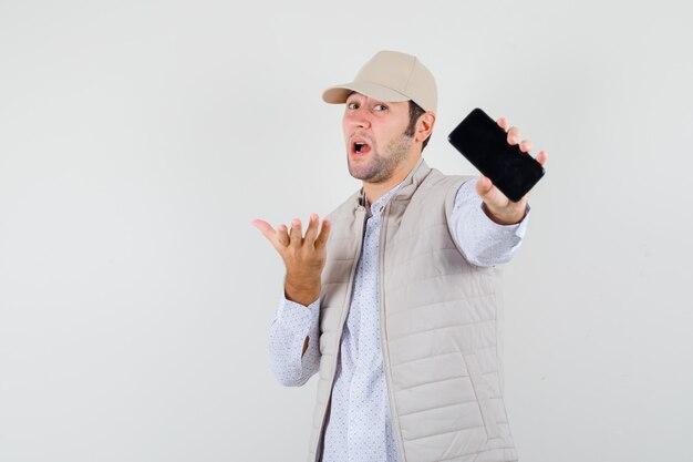 Молодой человек держит телефон в одной руке в бежевой куртке и кепке и выглядит оптимистично, вид спереди.