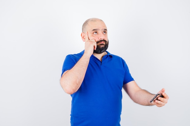 Free photo young man holding phone while looking up in blue shirt and looking pensive , front view.