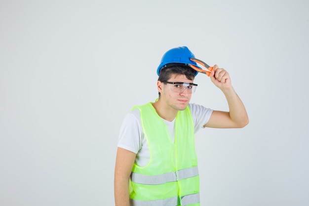 Young man holding pincers near head in construction uniform and looking confident , front view.