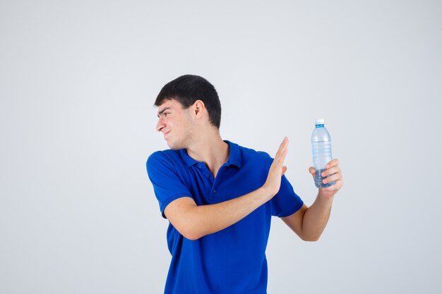 Young man holding plastic bottle, showing stop gesture in t-shirt and looking irritated