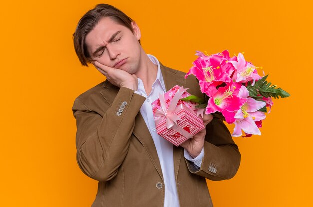 Молодой человек с подарком и букетом цветов выглядит усталым и скучающим и собирается поздравить с международным женским днем, стоя над оранжевой стеной