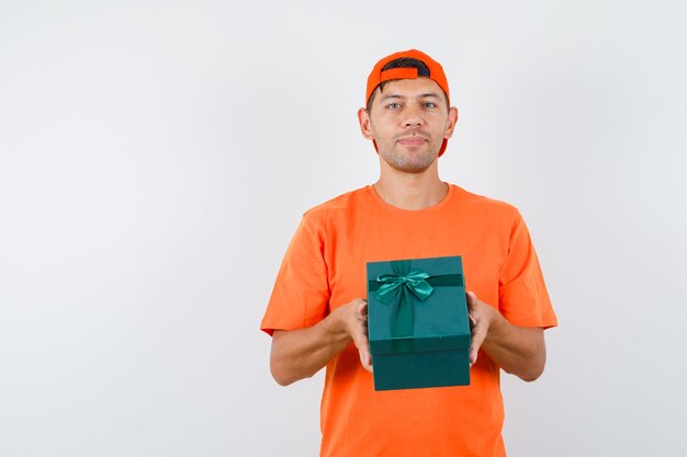 Молодой человек держит подарочную коробку в оранжевой футболке и кепке и выглядит уверенно