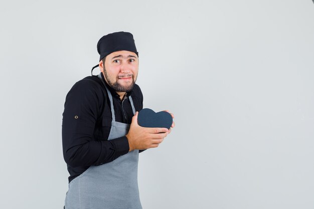 Young man holding present box in shirt, apron and looking agitated , front view.