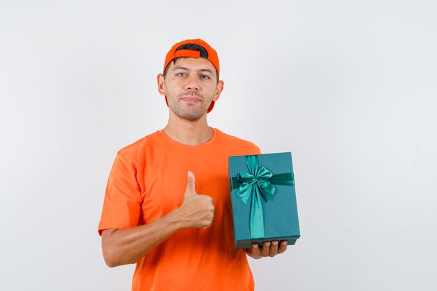 Young man holding present box with thumb up in orange t-shirt and cap and looking pleased