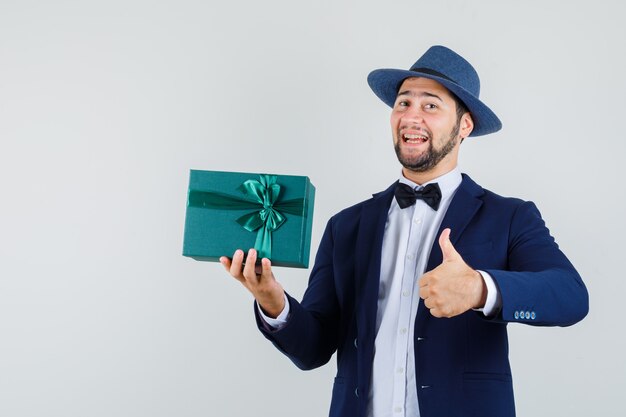 Young man holding present box with thumb up in suit, hat and looking glad. front view.